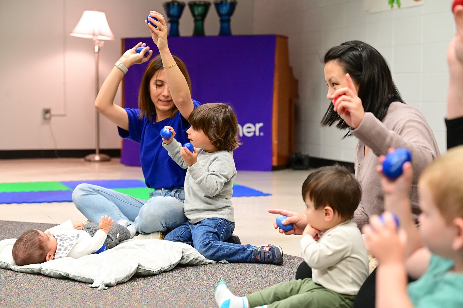 A young baby sits low on the floor among older toddlers and a teacher, watching the bigger kids who already know every song — the mixed-ages room at work.