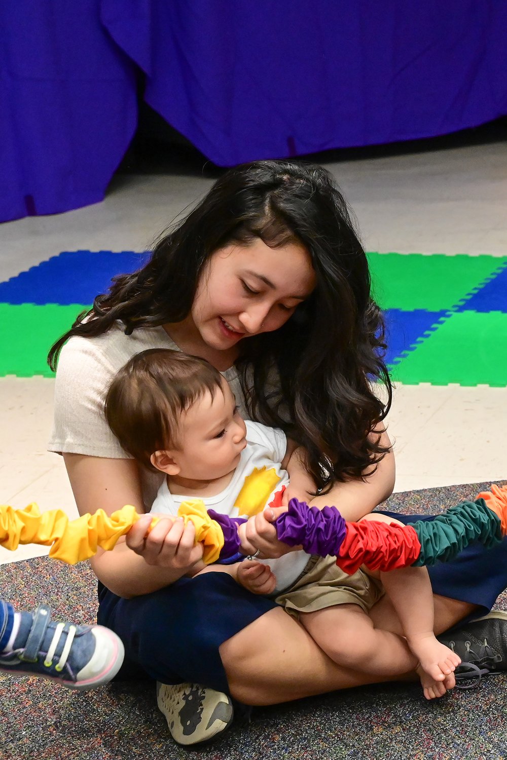 A grown-up holds a baby close on her lap as the two of them stretch a colorful band together — the easy warmth of a room your child knows by heart.