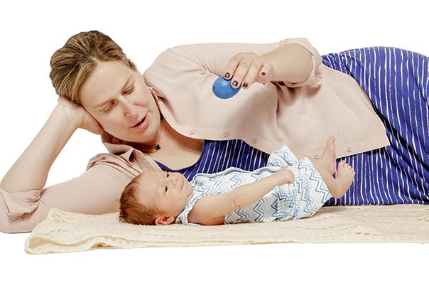 A mother lies on the floor beside her young baby during class, the two of them quiet and close while the music plays around them.