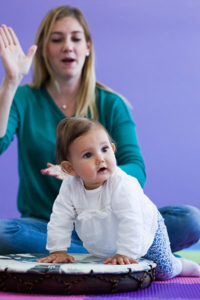 A baby lifts her head, bright and alert, as the grown-up behind her sings with a raised hand — turning toward the music the moment it begins.