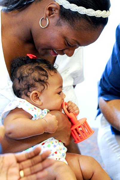A mother nuzzles her baby close as the baby grips a bright red shaker and brings it toward her mouth — small fingers just discovering that holding on makes the sound.