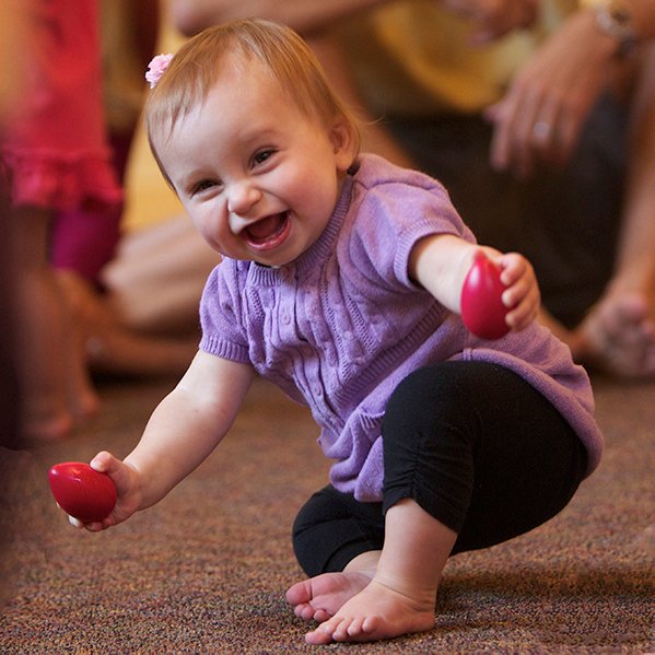 A baby crawls across the floor with a red egg shaker already in hand and a huge grin, moving with purpose toward the rest of the instruments.