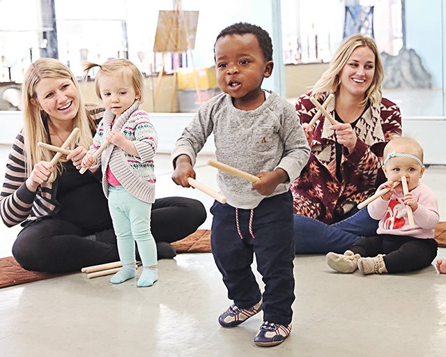 A toddler stands in the middle of the circle with rhythm sticks in hand, calling out mid-song while grown-ups play along on either side — a child who has decided this room is his.