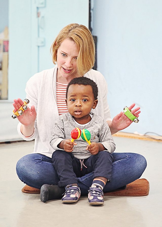 A toddler holds a maraca in each hand on his grown-up's lap while she plays the jingle bells behind him — the two of them making the music together now.