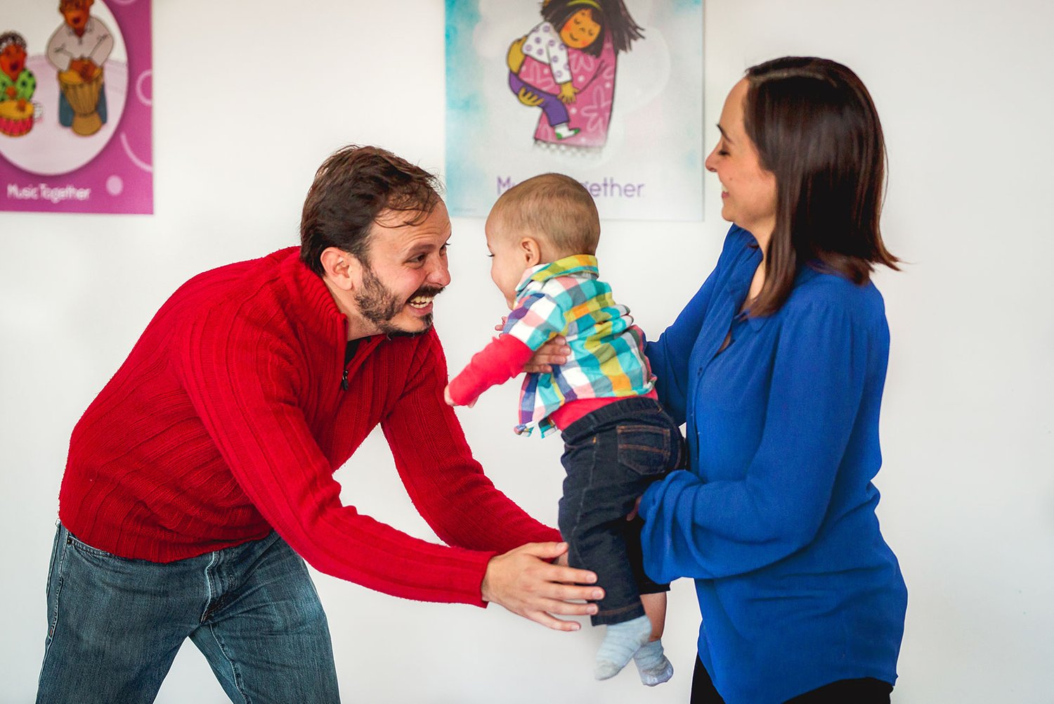 A baby stands supported in one parent's hands and reaches out toward the other — the wobbly, delighted discovery of being up on both feet in the middle of class.