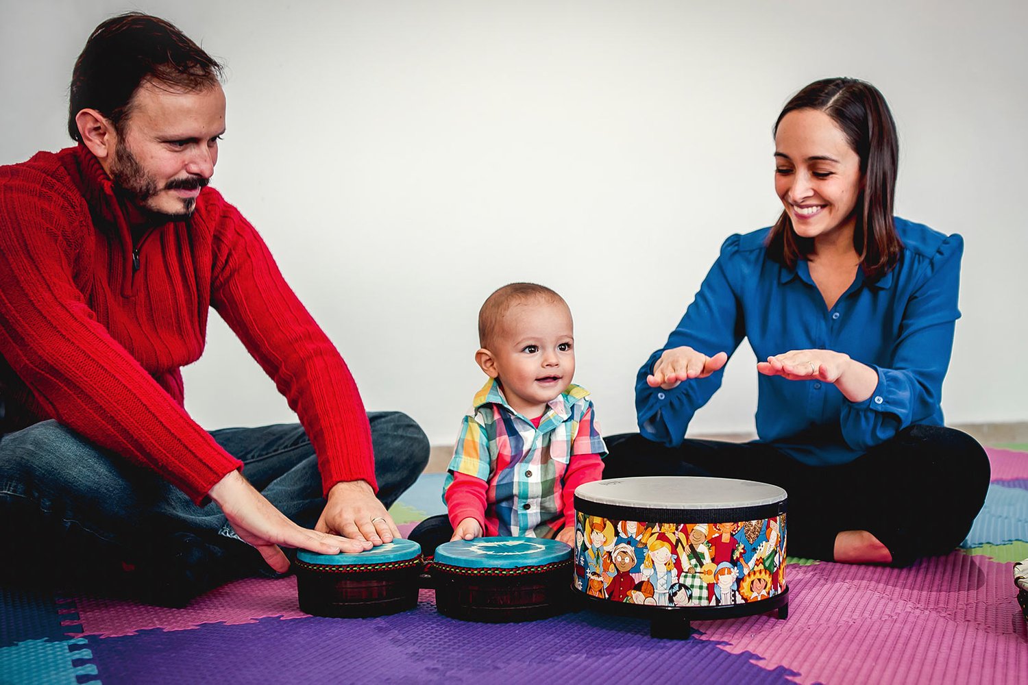 A baby sits between two grown-ups at the big gathering drums, every set of hands tapping the drumheads together — cause, effect, and shared rhythm in one moment.