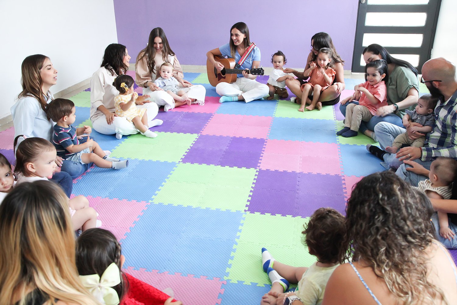 A full circle of families — babies, toddlers, and caregivers together in one room, a guitar in the middle — the community built one class at a time over a year.