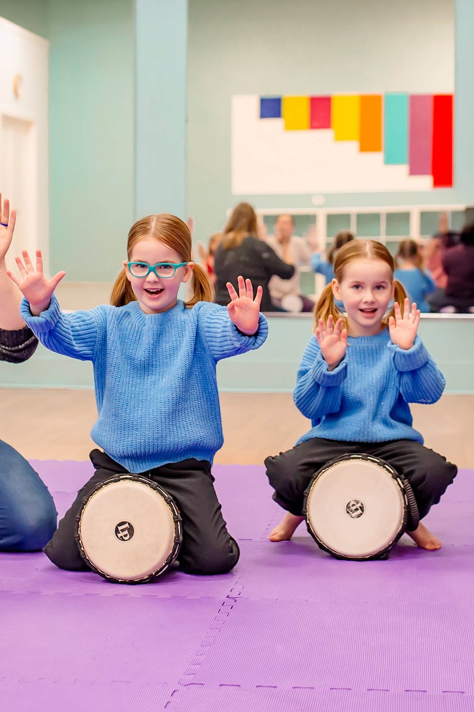 Two preschool-age children sitting with djembe drums, hands raised and smiling